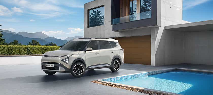 front three-quarter view of a Kia Carens Clavis parked outside a modern house with a pool