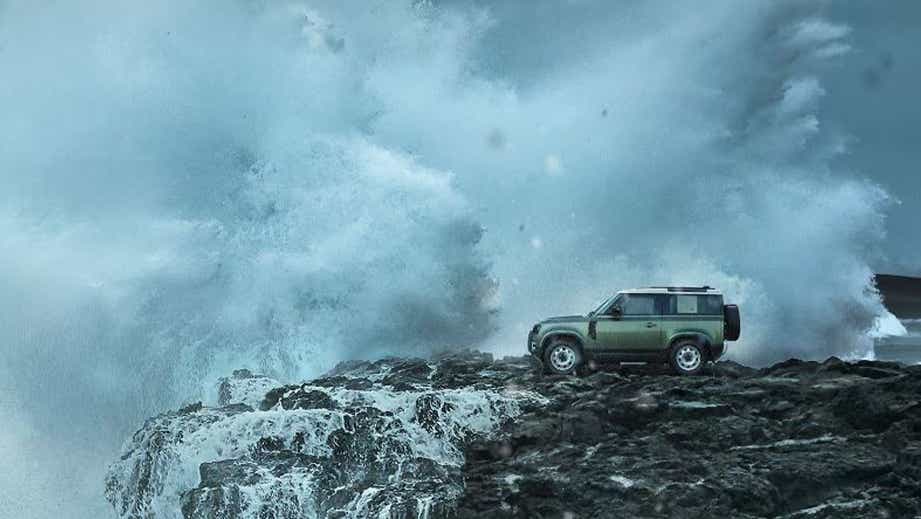 side view of a Land Rover Defender on rocky coastline with crashing waves