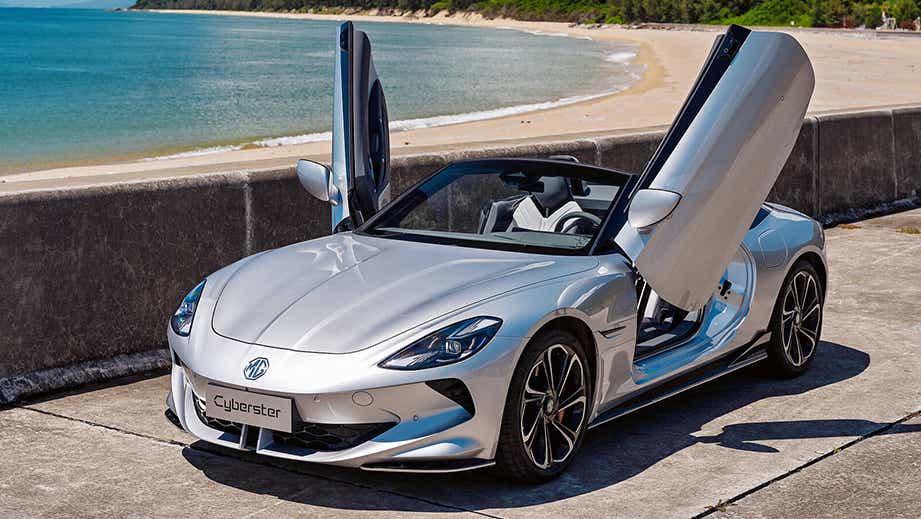 Front three-quarter view of an MG Cyberster convertible with doors open on a beachfront